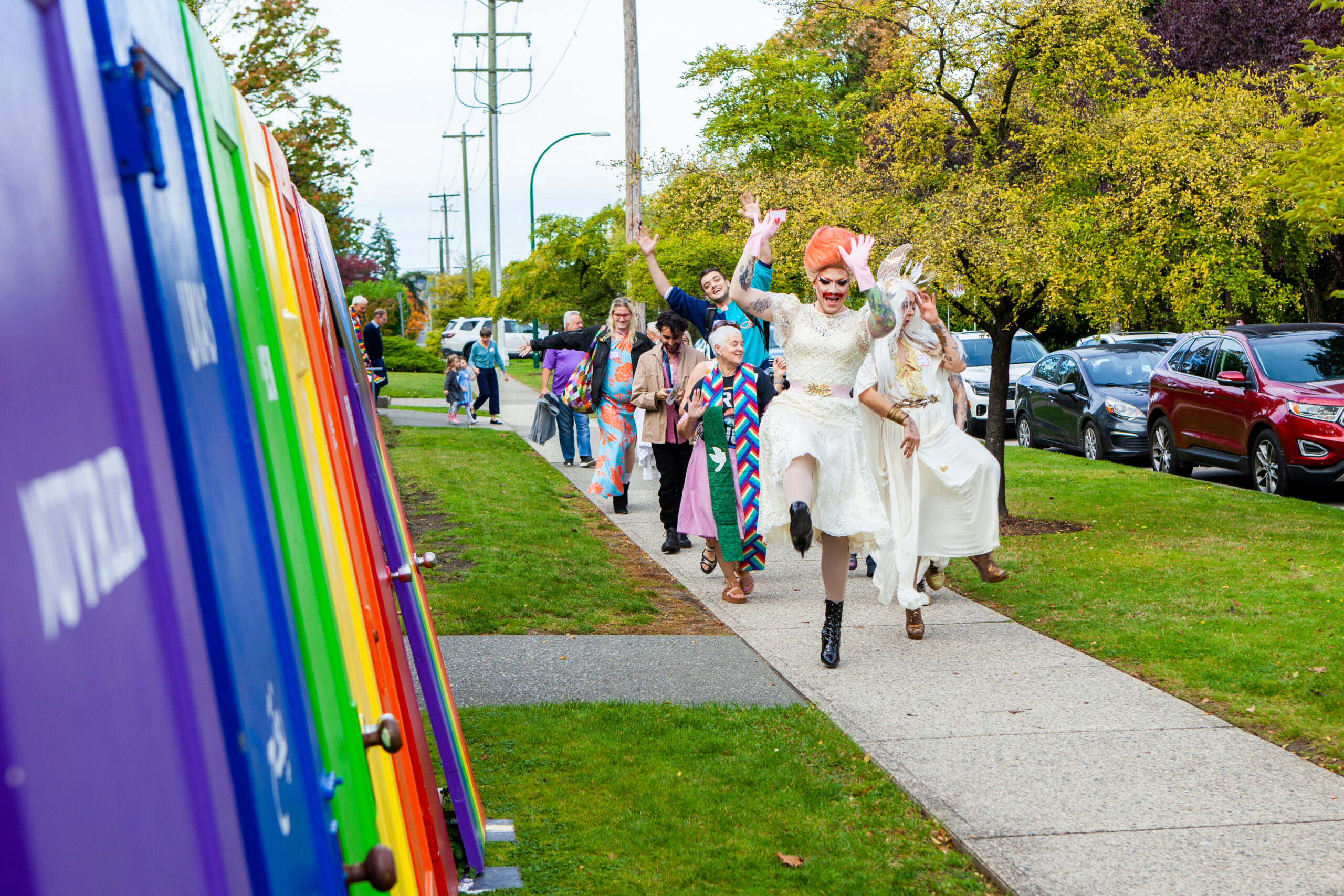 A brightly-dressed group of people, led by drag queen liturgists, parades excitedly down the sidewalk. To the side are several rainbow-coloured doors.